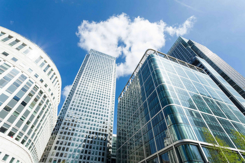 A view of modern glass skyscrapers and an office building under a bright blue sky with scattered clouds.