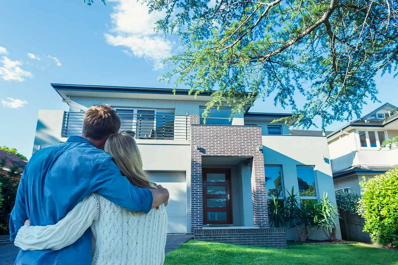 A couple stands arm-in-arm, looking at a modern two-story house.