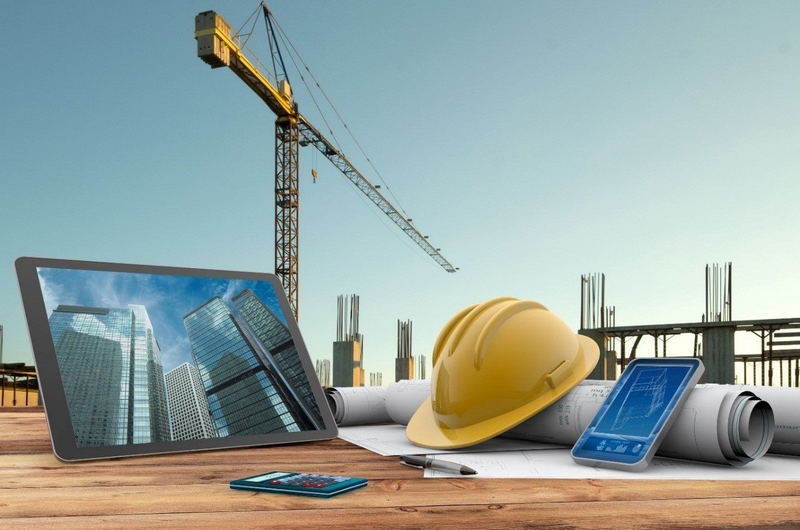 Construction scene with a crane, blueprints, a hard hat, and digital devices on a wooden table.