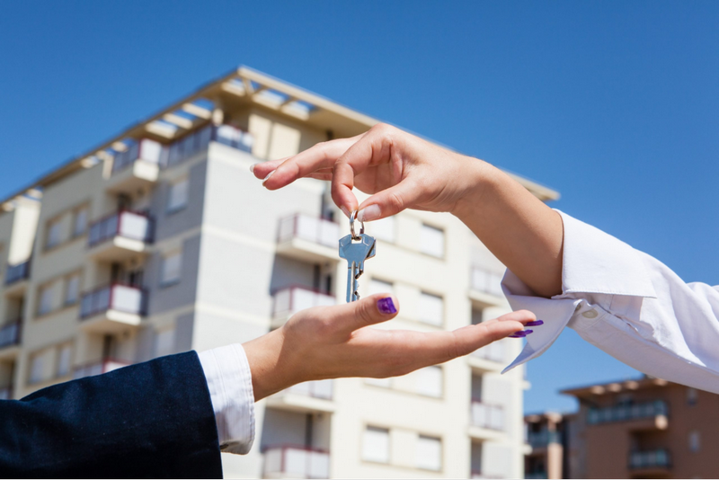 A hand is giving a key to another hand in front of an apartment building.